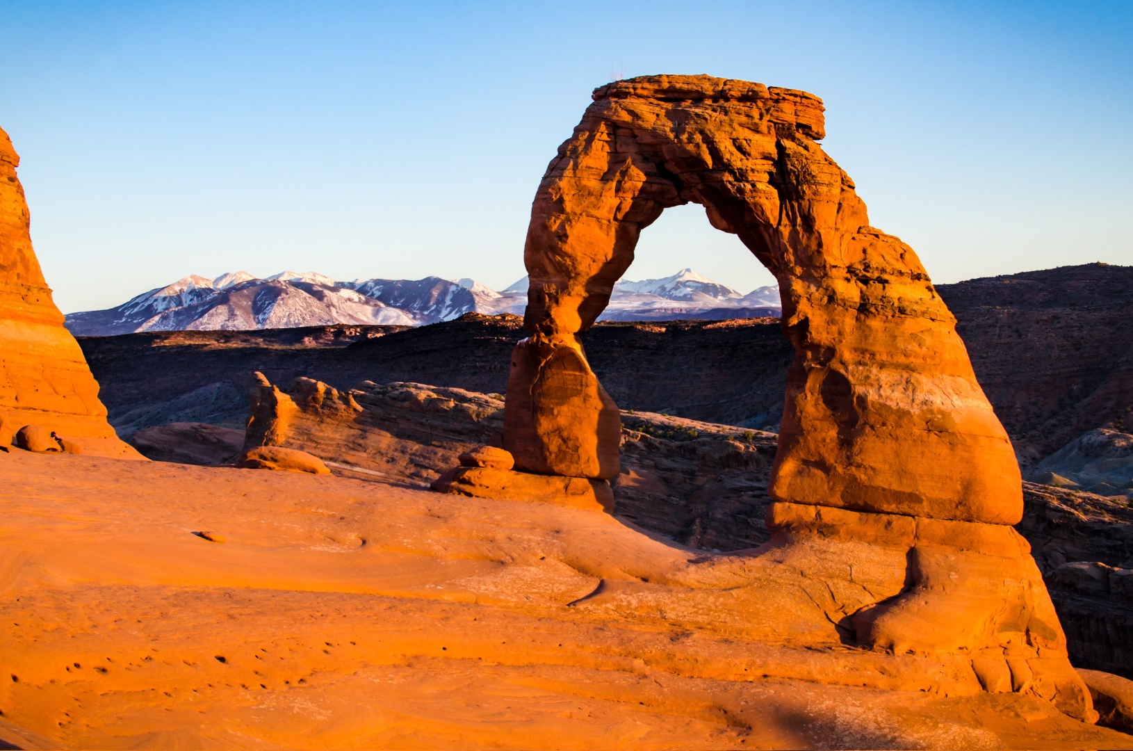 Delicate Arch glowing orange at sunset with snow-capped mountains beyond