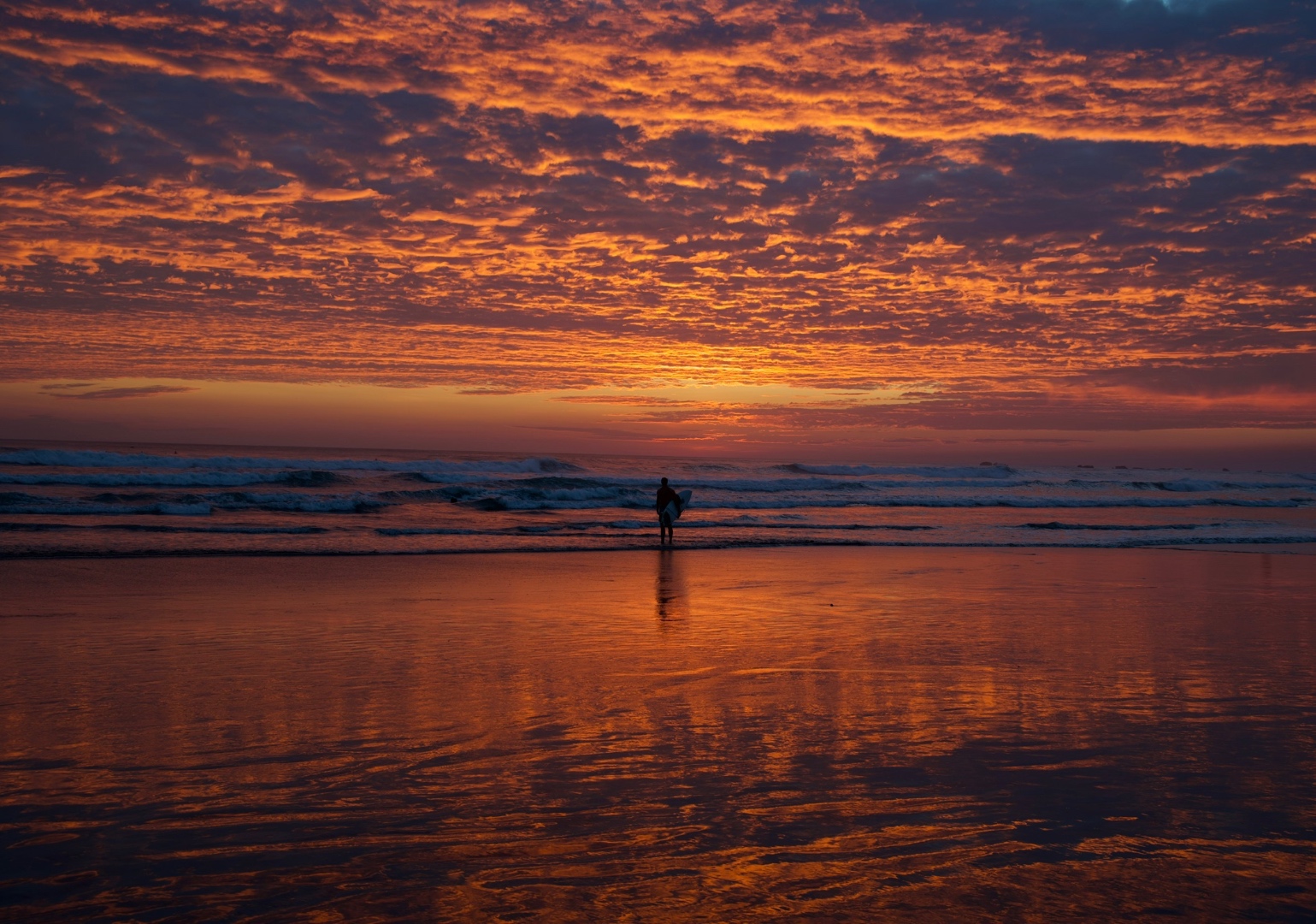 Silhouette of a surfer walking along a beach at a blazing orange sunset